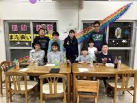 全体の集合写真。テラス側に二列に並んで撮影をした。正面左側の窓には「お別れ会、また会いましょう」のプラカードが窓に貼ってある。右側の窓の上部から左側の窓の一番下の左端に、柔らかい紙輪をつなげて作った虹が飾ってある。全長は約５メートル、色は上からピンク、赤、黄色、黄緑、緑、水色、青、紫。